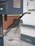 A close-up view of a green gas pump nozzle attached to a fuel dispenser. The background consists of a tiled wall and a concrete surface, revealing part of an industrial or service station environment.