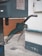 A close-up view of a green gas pump nozzle attached to a fuel dispenser. The background consists of a tiled wall and a concrete surface, revealing part of an industrial or service station environment.