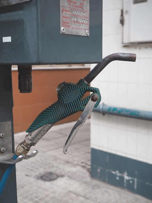 A close-up view of a green gas pump nozzle attached to a fuel dispenser. The background consists of a tiled wall and a concrete surface, revealing part of an industrial or service station environment.