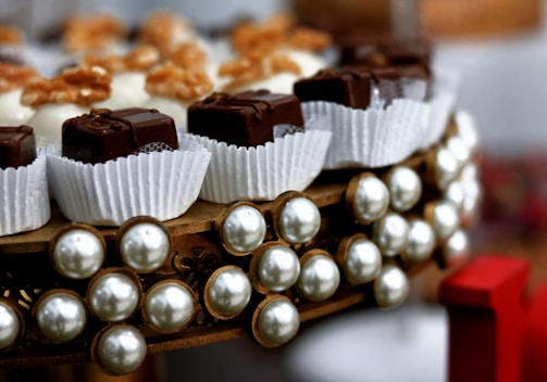 An arrangement of chocolate pieces wrapped in white paper cups is set on a decorative circular platter adorned with pearl-like beads. The chocolates are neatly aligned, and some are topped with nuts. The composition is elegant, with a focus on luxurious presentation.