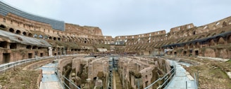A panoramic view of an ancient amphitheater with large, arched openings and tiered stone seating. The arena floor is exposed, showing subterranean passageways and structures. Restoration work is evident in some areas, with scaffolding and railings present.