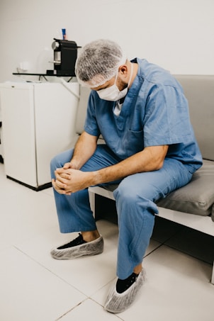 A person in medical scrubs, hairnet, and mask sits on a bench in a clinical setting, appearing contemplative or tired. The surroundings include a small refrigerator and a coffee maker, suggesting a break room in a healthcare environment.
