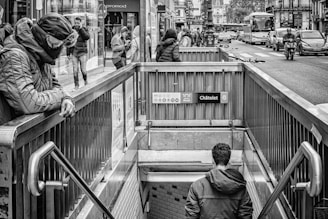 A busy urban street features people walking above and descending into a subway entrance marked with Châtelet signage. Vehicles, including buses and motorbikes, are visible on the street. The scene is in black and white, creating a classic city atmosphere.
