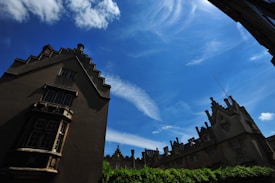 An architectural scene with historic buildings featuring ornate gabled roofs and numerous windows. The buildings are silhouetted against a bright blue sky with wispy, white clouds. A line of greenery borders the structures at the bottom.