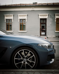 A parked luxury car is positioned on a street, showcasing its sleek lines and prominent front wheel. The background features a building with decorative elements around the windows, painted in muted colors, emphasizing the car's modern design.