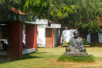 A statue depicting a seated figure in a meditative pose is situated in a grassy area surrounded by bountiful greenery. The scene includes overhanging tree branches and a building made of red brick with a tiled roof. The outdoor setting suggests a tranquil and peaceful atmosphere.