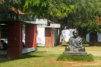 A statue depicting a seated figure in a meditative pose is situated in a grassy area surrounded by bountiful greenery. The scene includes overhanging tree branches and a building made of red brick with a tiled roof. The outdoor setting suggests a tranquil and peaceful atmosphere.