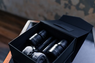 A neatly arranged set of black skincare bottles and jars inside an elegant black box. The box also contains rolled-up grey towels. The setting includes a wooden surface and a blurred background, creating a luxurious atmosphere.