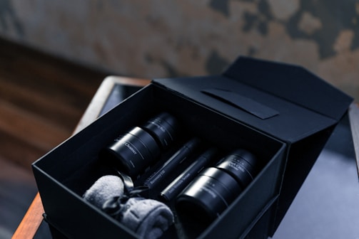 A neatly arranged set of black skincare bottles and jars inside an elegant black box. The box also contains rolled-up grey towels. The setting includes a wooden surface and a blurred background, creating a luxurious atmosphere.