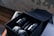 A neatly arranged set of black skincare bottles and jars inside an elegant black box. The box also contains rolled-up grey towels. The setting includes a wooden surface and a blurred background, creating a luxurious atmosphere.