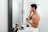 A shirtless man in a bathroom applies cream to his face while looking in a mirror. He holds a small jar in one hand and is surrounded by various grooming products on the sink. The bathroom is modern and minimalist, with a clean white color scheme.