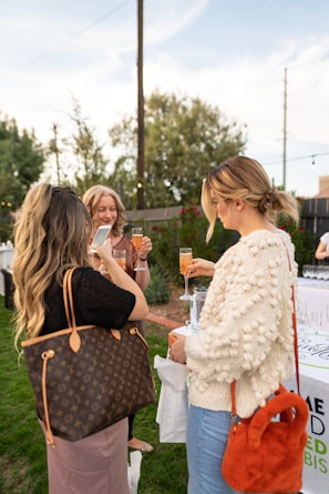 A group of women are socializing outdoors, holding drinks and talking. One is using a smartphone while others are smiling. They are near a table with empty glasses and in a garden setting.