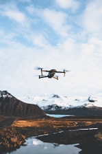 A drone is flying in the sky over a picturesque landscape. Snow-capped mountains stand in the background, and a body of water reflects the scenery. The foreground features a rugged terrain with patches of grass and rock.