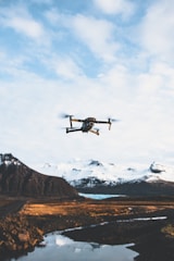 A drone is flying in the sky over a picturesque landscape. Snow-capped mountains stand in the background, and a body of water reflects the scenery. The foreground features a rugged terrain with patches of grass and rock.