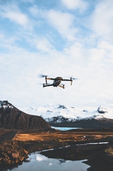 A drone is flying in the sky over a picturesque landscape. Snow-capped mountains stand in the background, and a body of water reflects the scenery. The foreground features a rugged terrain with patches of grass and rock.
