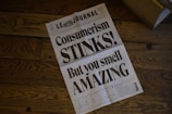 A newspaper lies open on a wooden floor, displaying a bold headline that critiques consumerism. The design is minimalist, with large black text over a white background, and smaller text at the bottom that resembles typical newspaper sections.
