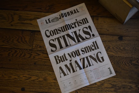 A newspaper lies open on a wooden floor, displaying a bold headline that critiques consumerism. The design is minimalist, with large black text over a white background, and smaller text at the bottom that resembles typical newspaper sections.