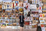 A person stands in front of a diverse collection of paintings displayed on an outdoor wall. The artworks vary in style, depicting scenes such as cityscapes, people, and still lifes with flowers. The person appears to be observing the art closely.