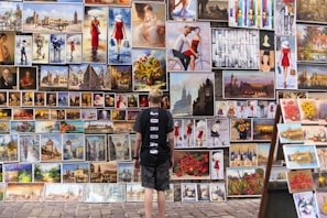 A person stands in front of a diverse collection of paintings displayed on an outdoor wall. The artworks vary in style, depicting scenes such as cityscapes, people, and still lifes with flowers. The person appears to be observing the art closely.