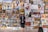 A person stands in front of a diverse collection of paintings displayed on an outdoor wall. The artworks vary in style, depicting scenes such as cityscapes, people, and still lifes with flowers. The person appears to be observing the art closely.
