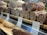 An assortment of salami displayed on wooden shelves in a deli or supermarket. The foreground shows several types of salami, including Calabrese and Varzi, all sliced to reveal their marbled interior. Each type is labeled with a small sign indicating its name and price.