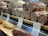 An assortment of salami displayed on wooden shelves in a deli or supermarket. The foreground shows several types of salami, including Calabrese and Varzi, all sliced to reveal their marbled interior. Each type is labeled with a small sign indicating its name and price.