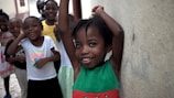 A group of smiling children stands together in a casual setting. The child in the foreground is leaning against a wall with arms raised, wearing a green and red dress. The children in the background are dressed in colorful clothing, and everyone appears cheerful and playful.