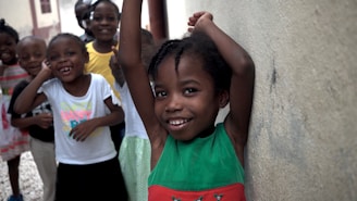 A group of smiling children stands together in a casual setting. The child in the foreground is leaning against a wall with arms raised, wearing a green and red dress. The children in the background are dressed in colorful clothing, and everyone appears cheerful and playful.