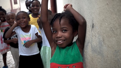 A group of smiling children stands together in a casual setting. The child in the foreground is leaning against a wall with arms raised, wearing a green and red dress. The children in the background are dressed in colorful clothing, and everyone appears cheerful and playful.