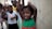 A group of smiling children stands together in a casual setting. The child in the foreground is leaning against a wall with arms raised, wearing a green and red dress. The children in the background are dressed in colorful clothing, and everyone appears cheerful and playful.