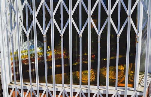 A metal gate with a crisscross pattern covers a window. Behind the window are several reflective disco balls, with gold and silver colors visible. Bright sunlight casts shadows and reflections, creating an interesting interplay of light and shadows.