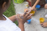 Children engaging in a creative activity outdoors, hands smudged with paint. Several containers with colorful paints are on the ground, and a child is seen mixing paint on their hands, focusing intently on the activity.