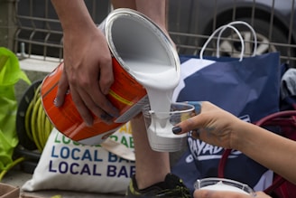 Two hands are seen pouring white liquid, possibly paint, from a large can into a smaller plastic cup. The background showcases various bags and a glimpse of a parked vehicle.