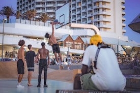 A group of young men are playing basketball outdoors. One player is mid-air, attempting to make a slam dunk. Others stand nearby, watching the action. In the background, there are modern buildings with a clear sky, palm trees, and spectators casually observing the game.