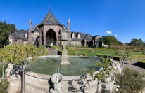 A large, elegant mansion with Gothic architectural features is situated in a landscaped garden. The home has steeply pitched roofs and numerous windows, with a grand entrance showcased by tall arches and columns. In front of the mansion, there is a circular stone fountain with water jets surrounded by manicured hedges and bushes.