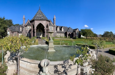 A large, elegant mansion with Gothic architectural features is situated in a landscaped garden. The home has steeply pitched roofs and numerous windows, with a grand entrance showcased by tall arches and columns. In front of the mansion, there is a circular stone fountain with water jets surrounded by manicured hedges and bushes.