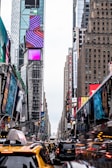 A bustling urban street scene featuring tall buildings with digital advertisements and billboards. The foreground includes yellow taxis and various vehicles in heavy traffic. Pedestrians navigate the crowded sidewalks by the colorful and vibrant signage.