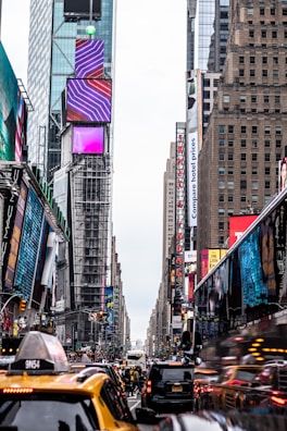 A bustling urban street scene featuring tall buildings with digital advertisements and billboards. The foreground includes yellow taxis and various vehicles in heavy traffic. Pedestrians navigate the crowded sidewalks by the colorful and vibrant signage.