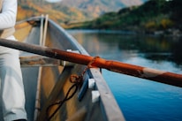 A canoe is floating on a calm river surrounded by lush greenery and distant mountains. An oar rests on the side, and part of a person wearing white clothing is visible inside the boat. The water reflects the natural landscape.