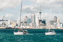 A scenic view of a harbor with modern skyscrapers in the background and two sailboats in the foreground. The city's skyline features a prominent needle-like tower surrounded by various high-rise buildings. The water is a rich, deep blue, and the sky is partly cloudy.