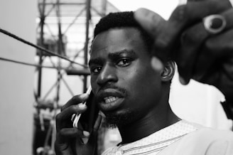 A person looking at the camera with a serious expression, holding a mobile phone to their ear. They are wearing a patterned shirt and a ring on their hand. The background includes a structure with ropes or cables.