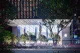 A modern, glass-covered building with a prominent logo on one side, set against a backdrop of lush green trees. The structure has a transparent facade that reveals an illuminated interior and people inside. The surrounding area includes more greenery and a road in the foreground with light trails suggesting moving vehicles.