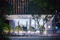 A modern, glass-covered building with a prominent logo on one side, set against a backdrop of lush green trees. The structure has a transparent facade that reveals an illuminated interior and people inside. The surrounding area includes more greenery and a road in the foreground with light trails suggesting moving vehicles.