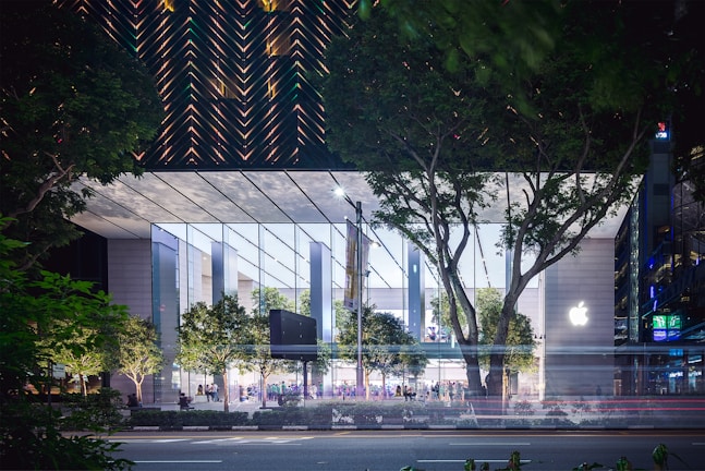 A modern, glass-covered building with a prominent logo on one side, set against a backdrop of lush green trees. The structure has a transparent facade that reveals an illuminated interior and people inside. The surrounding area includes more greenery and a road in the foreground with light trails suggesting moving vehicles.