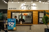 A counter with the name 'Janji Jiwa' above it, featuring a wooden panel design. Two people are behind the counter; they appear to work there. To the left, there are promotional posters, including one advertising a 10% cashback offer with GoPay. There are hanging lights and green plants adding to the decor.