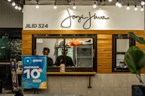 A counter with the name 'Janji Jiwa' above it, featuring a wooden panel design. Two people are behind the counter; they appear to work there. To the left, there are promotional posters, including one advertising a 10% cashback offer with GoPay. There are hanging lights and green plants adding to the decor.