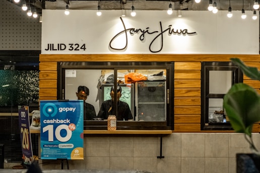 A counter with the name 'Janji Jiwa' above it, featuring a wooden panel design. Two people are behind the counter; they appear to work there. To the left, there are promotional posters, including one advertising a 10% cashback offer with GoPay. There are hanging lights and green plants adding to the decor.