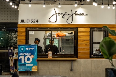 A counter with the name 'Janji Jiwa' above it, featuring a wooden panel design. Two people are behind the counter; they appear to work there. To the left, there are promotional posters, including one advertising a 10% cashback offer with GoPay. There are hanging lights and green plants adding to the decor.
