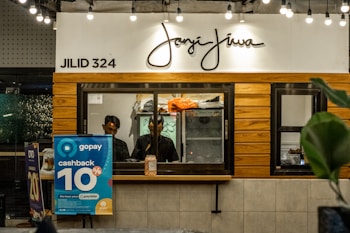 A counter with the name 'Janji Jiwa' above it, featuring a wooden panel design. Two people are behind the counter; they appear to work there. To the left, there are promotional posters, including one advertising a 10% cashback offer with GoPay. There are hanging lights and green plants adding to the decor.
