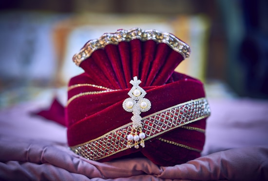 An ornate, deep red velvet turban adorned with intricate golden embroidery and embellishments, accented by a central decorative brooch featuring pearls and gem-like stones. The rich fabric and embellishments create a luxurious and culturally traditional appearance.